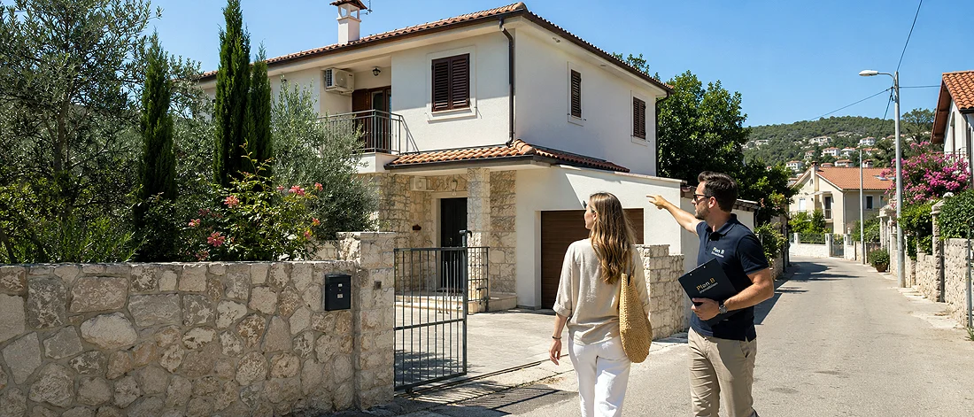 Haus in Ulcinj kaufen - Ein Mann und eine Frau stehen vor einem von einem Tor umgebenen Steinhaus mit Terrakottadach. Der Mann zeigt auf das Haus, während er ein Klemmbrett in der Hand hält. Die Straße ist von ähnlichen Häusern und Grünflächen gesäumt.