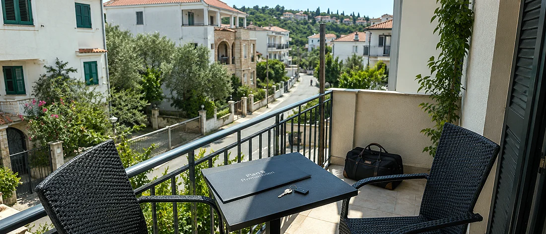 Ein kleiner Balkon mit zwei schwarzen Stühlen und einem Tisch, auf dem eine Mappe, ein Stift und ein Zimmerschlüssel liegen, bietet einen Blick auf eine ruhige Wohnstraße, die von Häusern und Grünflächen gesäumt ist. Eine schwarze Tasche steht auf dem Boden an der Wand.