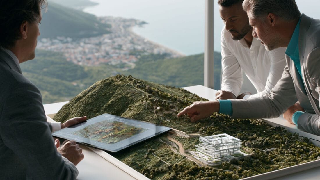 Drei Männer in Geschäftskleidung besprechen ein detailliertes Architekturmodell einer hügeligen Landschaft mit Gebäuden, Straßen und Grünflächen, während einer von ihnen ein Tablet benutzt. Durch das Fenster im Hintergrund ist eine Küstenstadt zu sehen.
