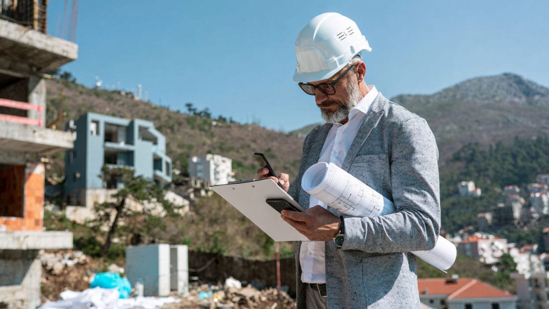 Objektprüfung - Mann mit Bauhelm prüft auf einer Baustelle Baupläne, Klemmbrett und Smartphone.
