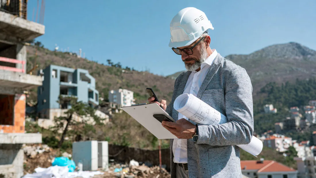 Objektprüfung - Mann mit Bauhelm prüft auf einer Baustelle Baupläne, Klemmbrett und Smartphone.