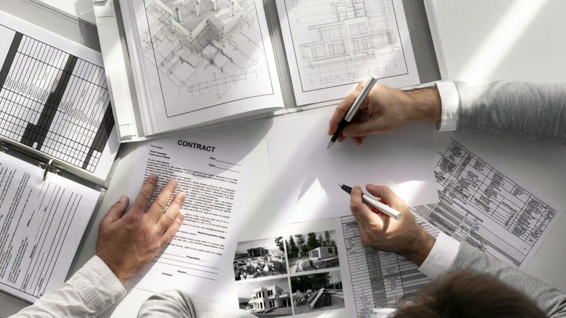 Risiken Immobilienkauf - Zwei Personen sitzen an einem Schreibtisch und prüfen Baupläne, Blaupausen, einen Vertrag und Fotos von Gebäuden. Die eine macht sich Notizen, während die andere die Unterlagen hält. Auf dem weißen Schreibtisch liegen Dokumente und technische Zeichnungen ausgebreitet.