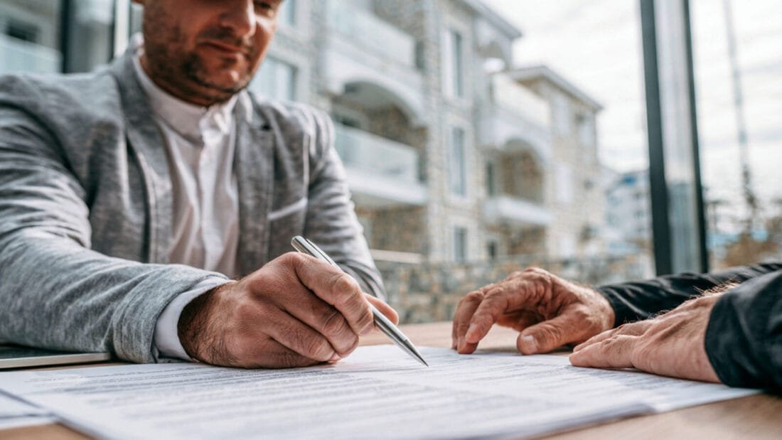 Zwei Personen sitzen an einem Tisch mit Dokumenten; eine Person unterschreibt, die andere zeigt auf das Papier. Im Hintergrund sind Fenster und Gebäude zu sehen.