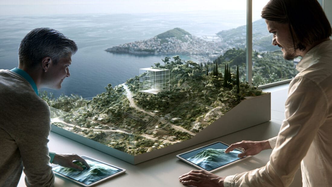 Zwei Personen stehen an einem Tisch, auf dem ein 3D-Architekturmodell einer hügeligen Landschaft steht. Beide nutzen ein Tablet, auf dem das digitale Gelände angezeigt wird. Durch die großen Fenster hinter ihnen sind ein Gewässer und eine Küstenstadt zu sehen.
