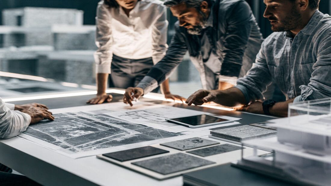 Vier Personen stehen um einen Tisch herum und betrachten Baupläne, Materialmuster und maßstabsgetreue Modelle. Der Schauplatz scheint ein modernes Büro oder Atelier mit großen Fenstern und zeitgemäßer Einrichtung zu sein.